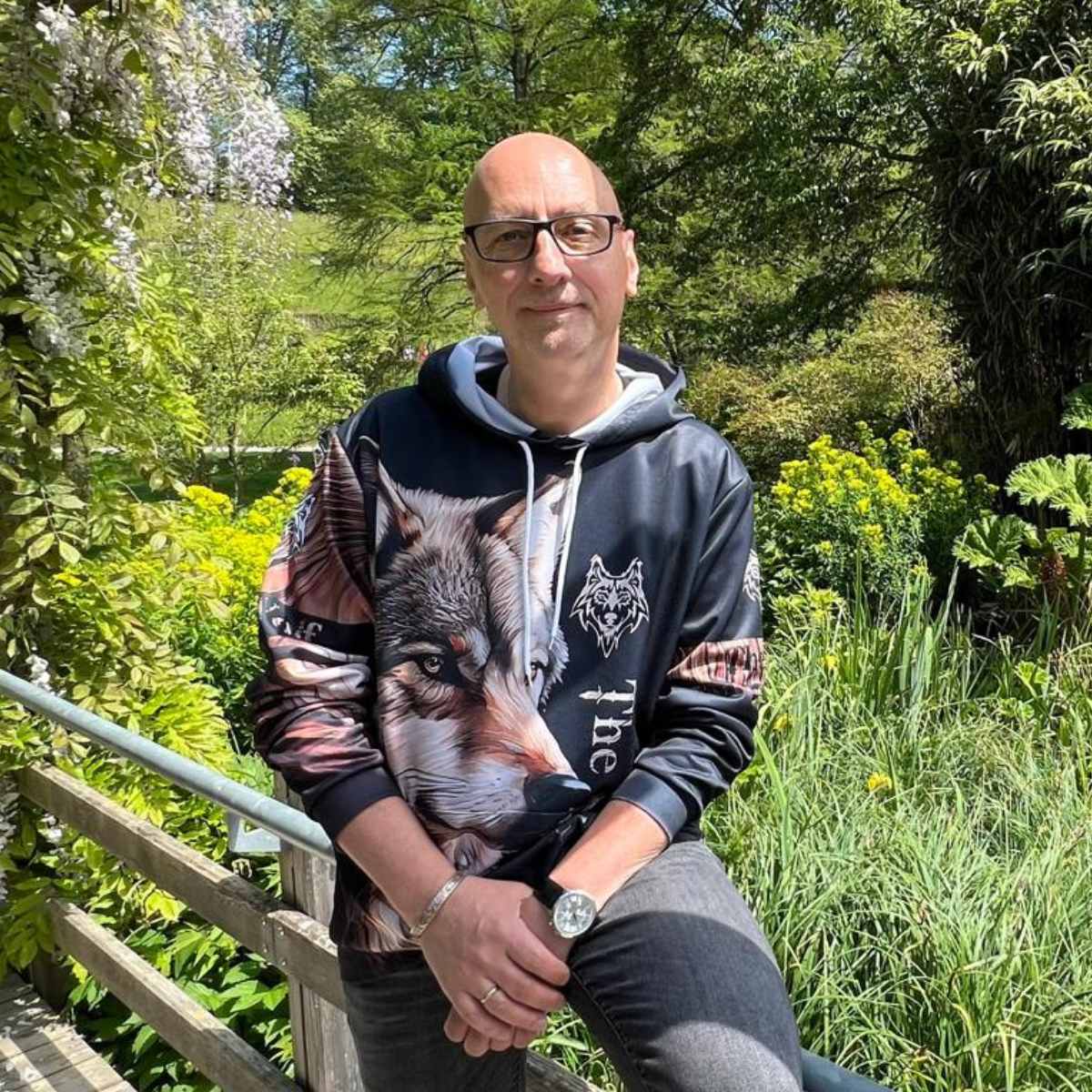 Porträt von Ralf Musto, systemischer Transformations-Coach und Hypnotherapeut, der entspannt auf einer Holzbrücke in einem sonnenbeschienenen Garten sitzt. Er trägt einen Hoodie mit Wolfsmotiv und blickt ruhig in die Kamera. Symbolbild für den Ausstieg aus dem Hamsterrad und die Rückverbindung zur eigenen Natur.